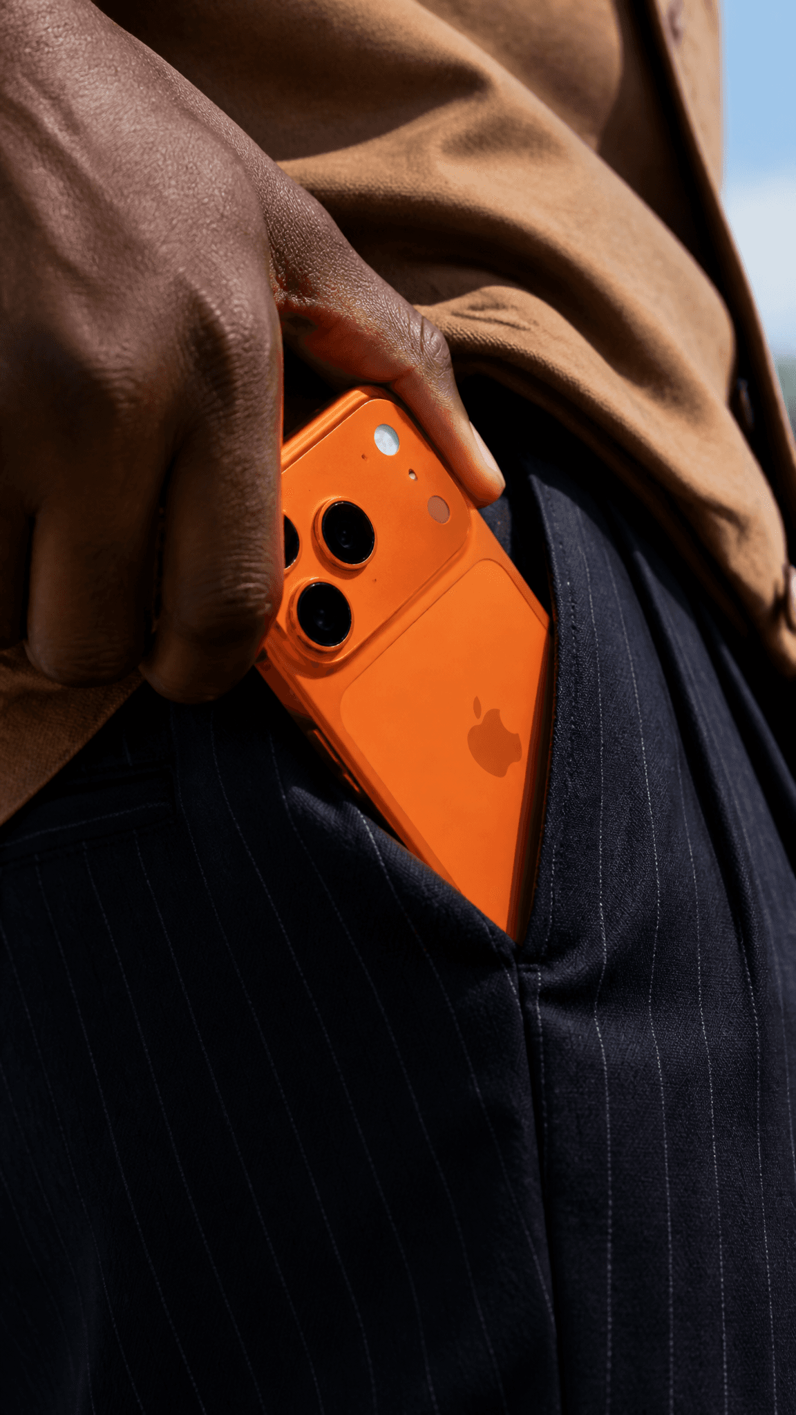 A close-up shot shows a person's dark-skinned hand, likely belonging to a Black male of indeterminate age, as it tucks an orange smartphone into the pocket of dark, pinstriped trousers. The smartphone, featuring a prominent triple-camera module and the Apple logo, is positioned diagonally within the pocket, with its top edge near the hand. The background is softly blurred, revealing a light brown fabric, possibly a jacket or shirt, and a hint of light blue sky. The lighting is soft and directional, casting subtle shadows and highlighting the textures of the clothing and the phone's surface. The overall atmosphere is clean, modern, and suggestive of an everyday, candid moment, with a focus on the vibrant color of the phone against the muted tones of the clothing. The perspective is slightly angled, emphasizing the diagonal line of the phone and the action of tucking it away.